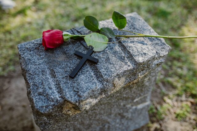 Cruce simplă de lemn pe un mormânt proaspăt, acoperit cu flori și coroane, într-un cimitir liniștit