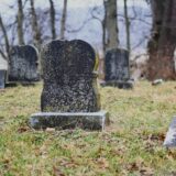 Monumente funerare din granit și marmură, aliniate într-un cimitir liniștit, înconjurate de copaci verzi